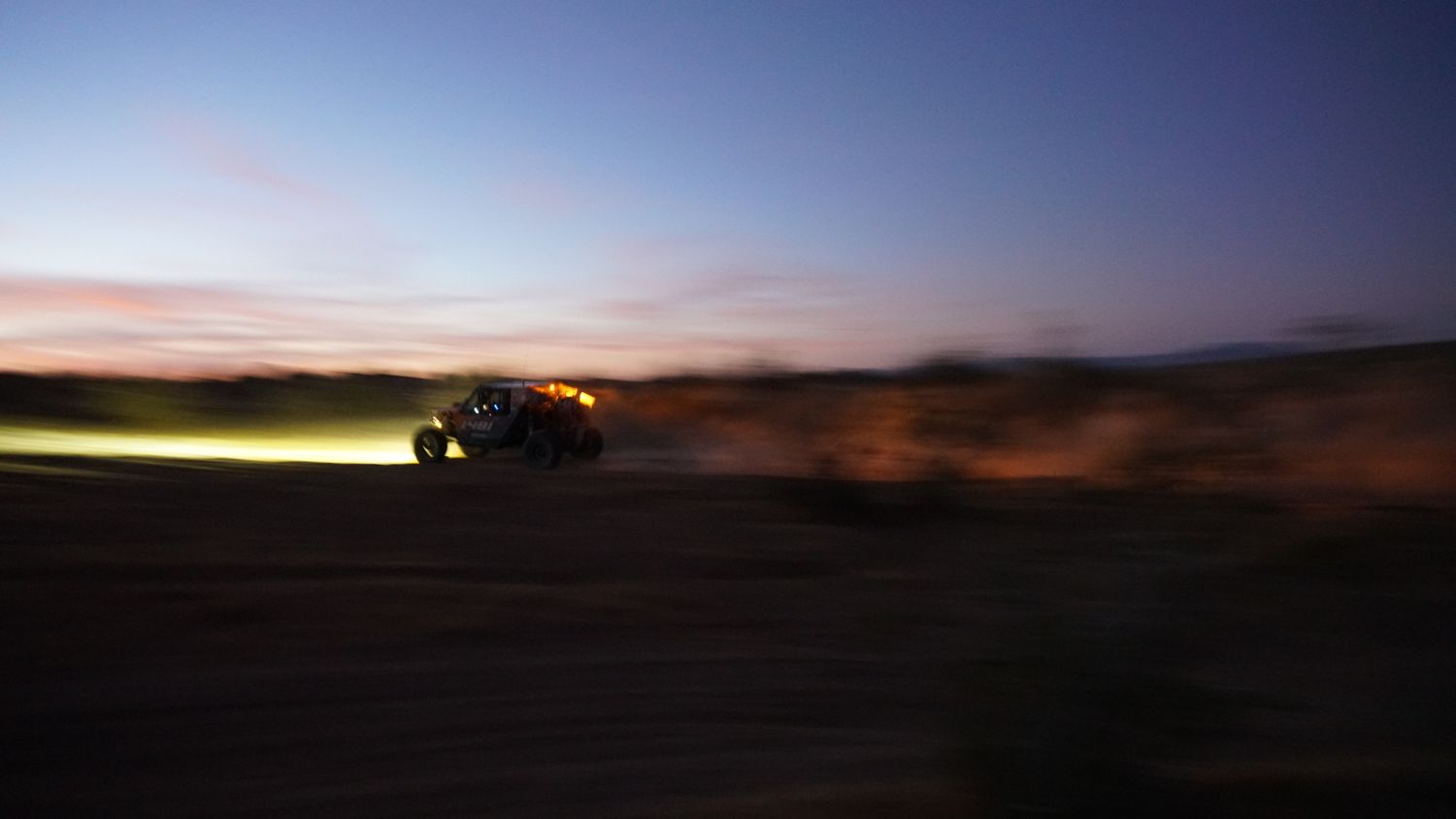 Off-road vehicle racing across desert terrain kicking up dust at dusk with glowing lights.