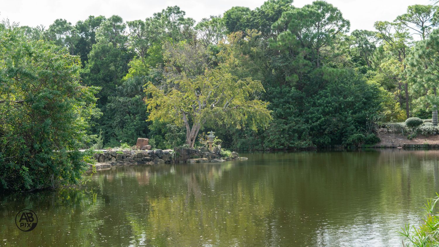 Tranquil pond reflecting lush green trees and a small rocky island in a peaceful landscape.