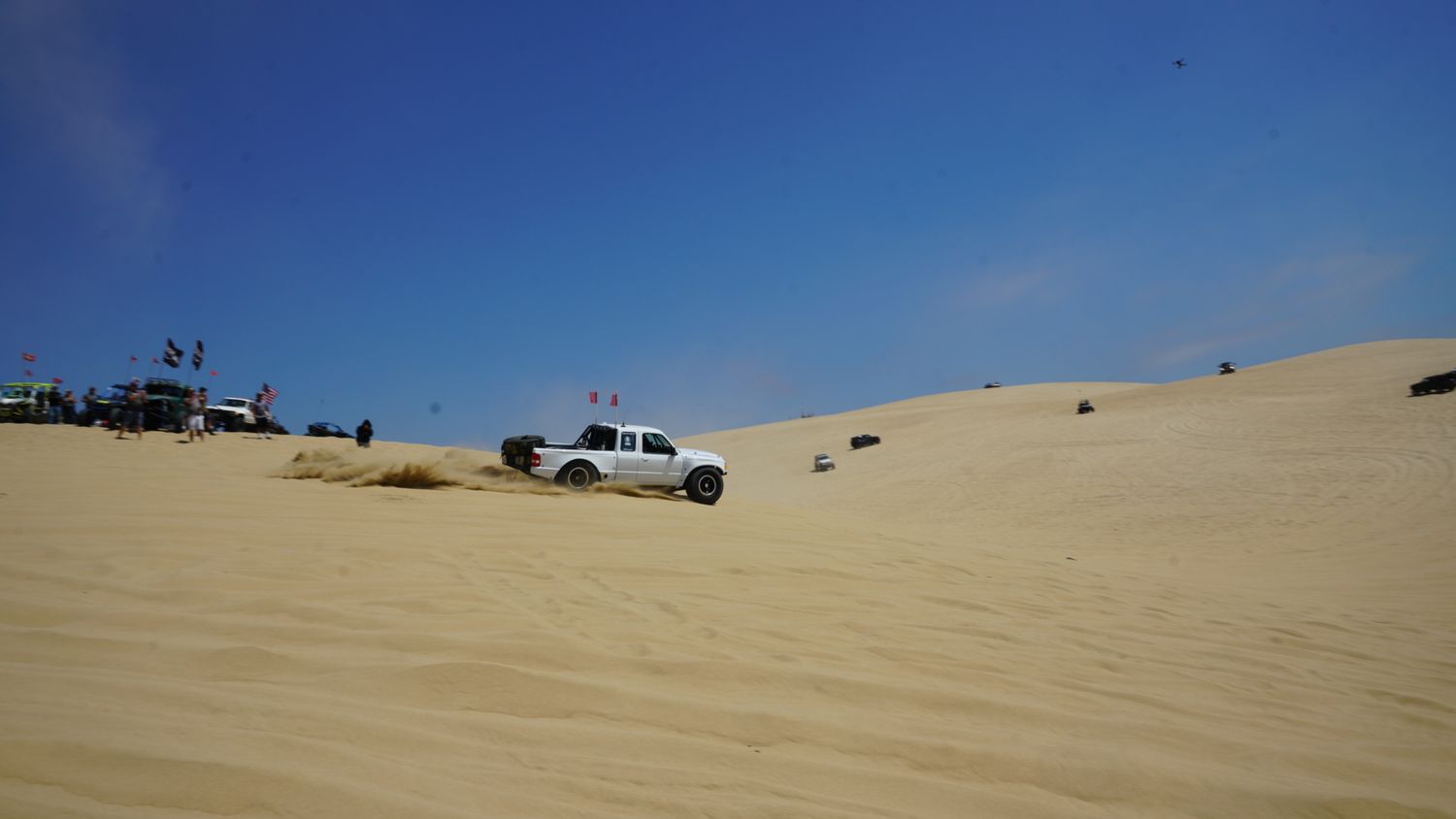 White off-road truck climbs a steep sand dune at an outdoor desert motorsport gathering.