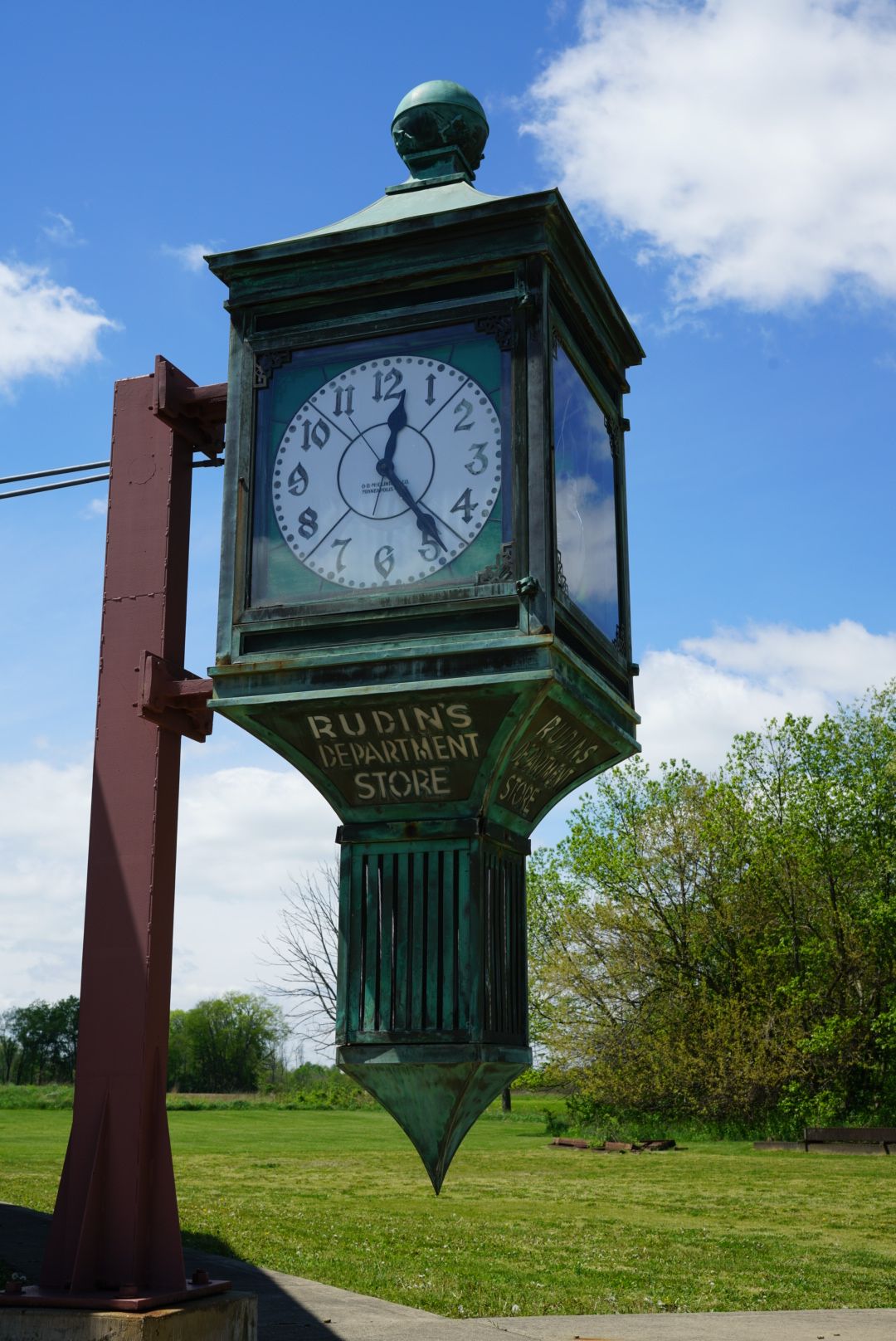 Antique green patina Rudins Department Store street clock mounted on a red metal post in a park.