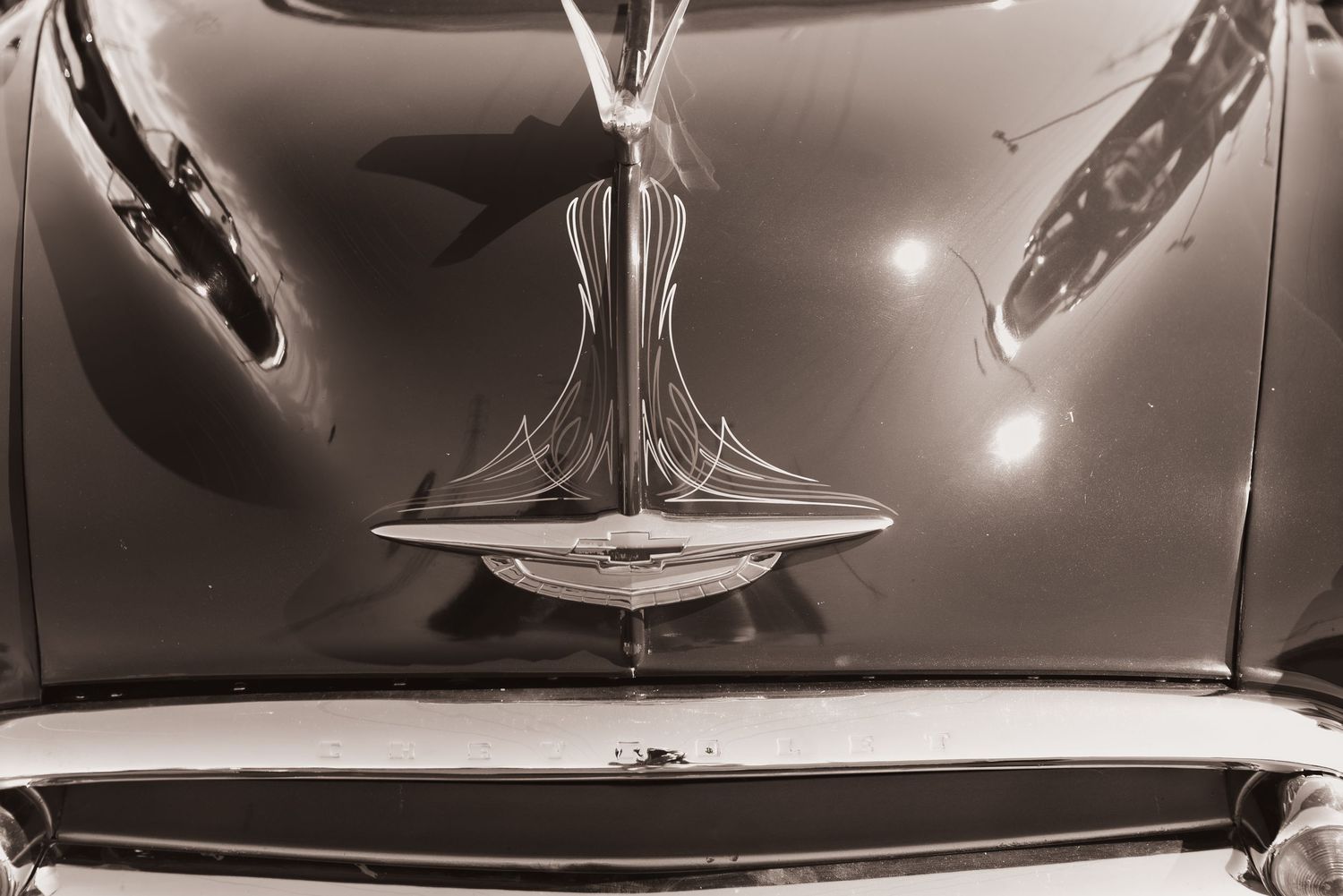 Sepia-toned close-up of a vintage classic car hood ornament and chrome grille detail.