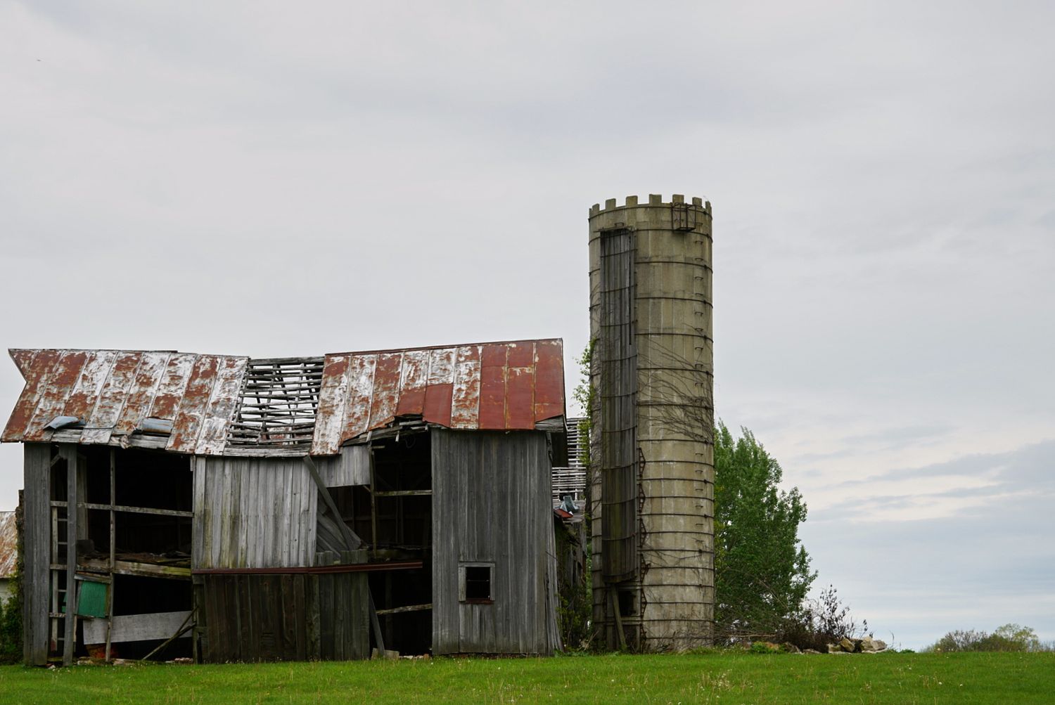 Abandoned weathered barn with rusted corrugated metal roof beside a stone silo on a green grassy rural property.