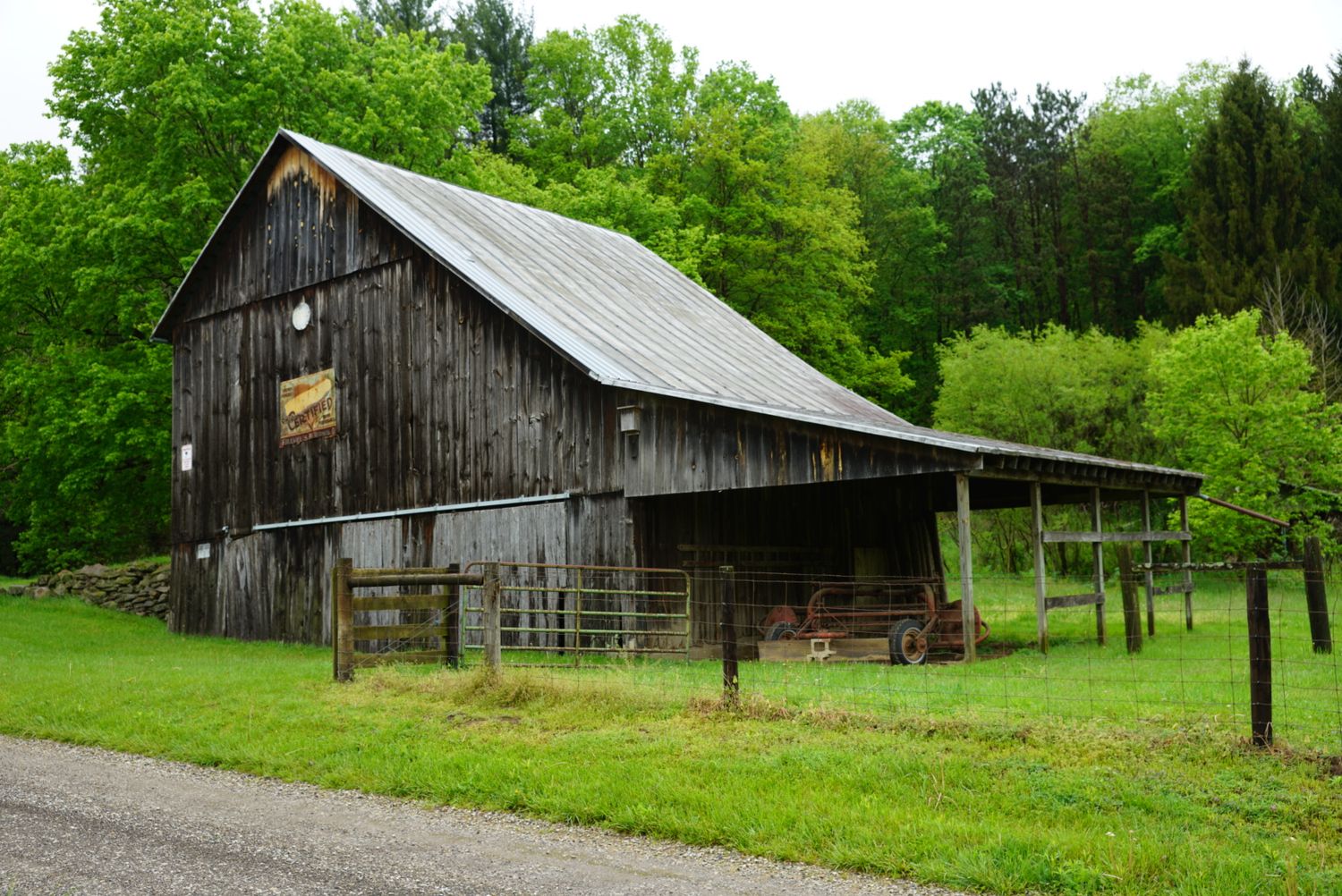 Rustic weathered wooden barn with metal roof surrounded by lush green trees on a rural farm property.