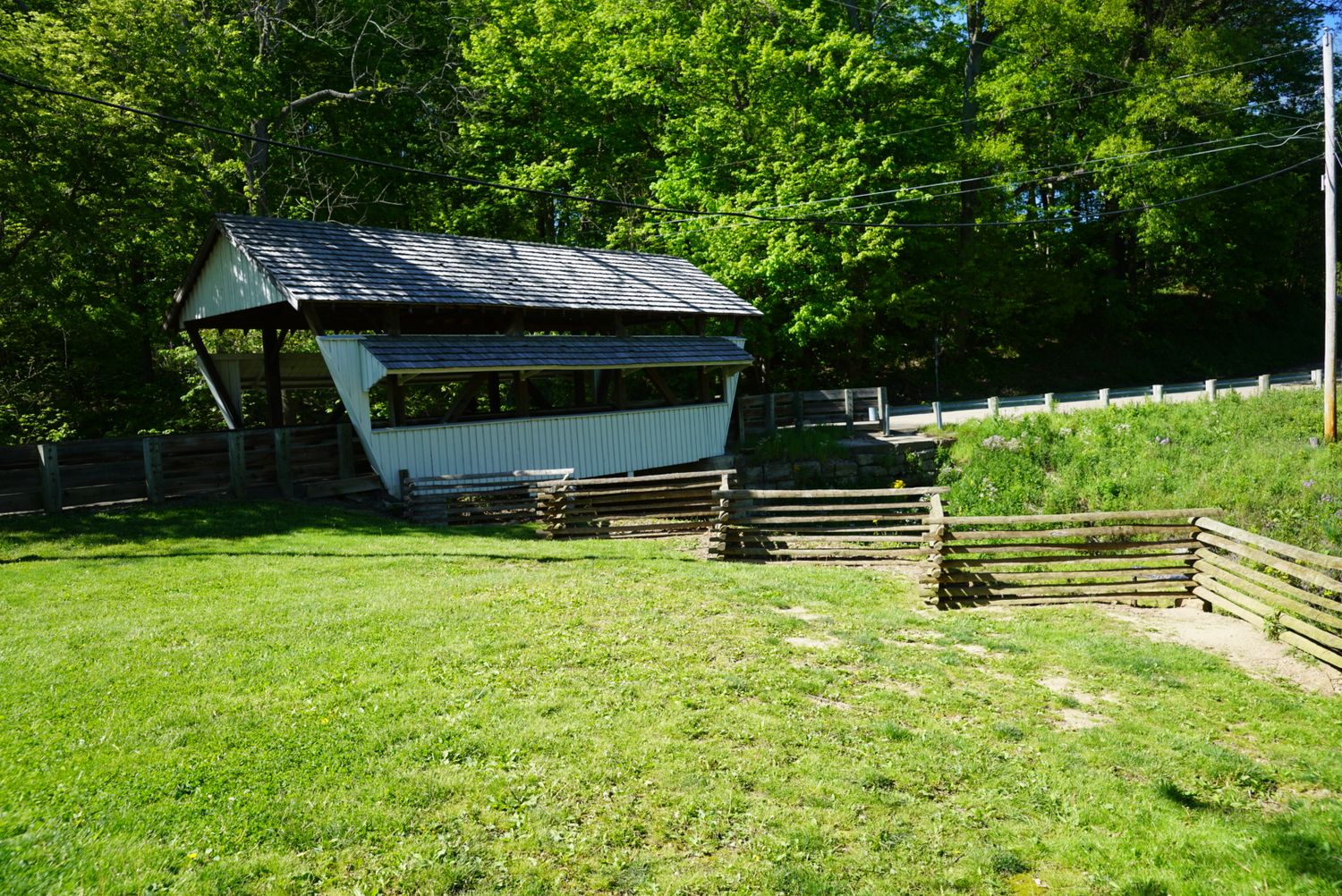 A historic covered bridge surrounded by lush green trees with a wooden split-rail fence in a rural countryside setting.
