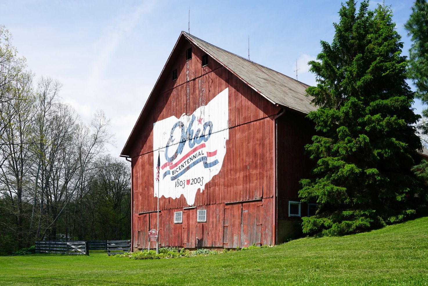 Red barn with Ohio bicentennial state outline mural painted on side in green countryside.