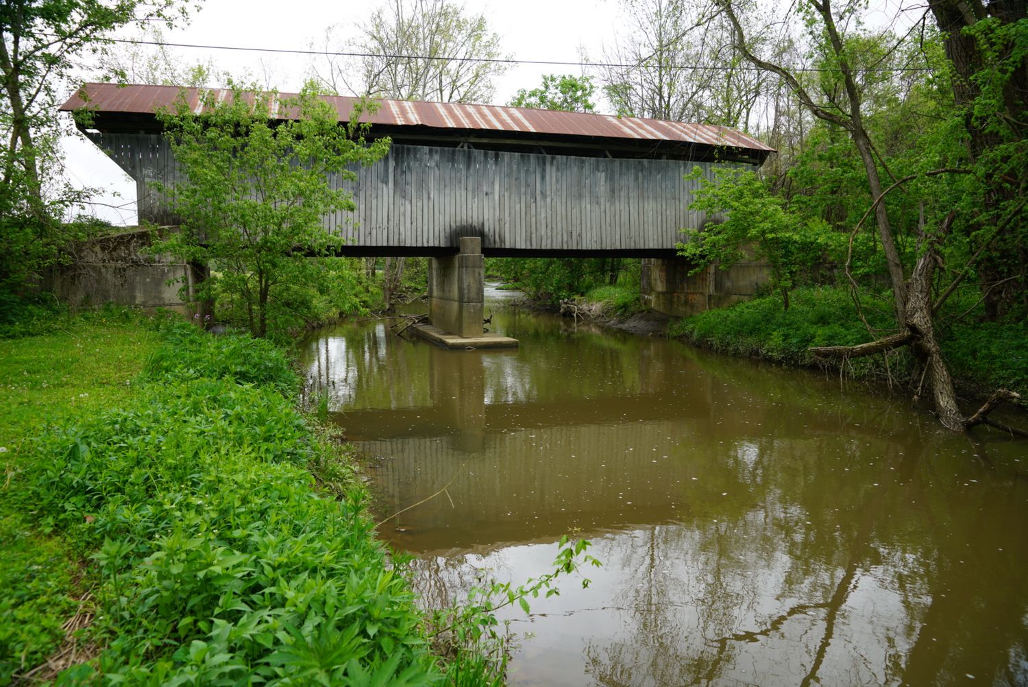 A historic wooden covered bridge reflects in calm water surrounded by lush green trees.