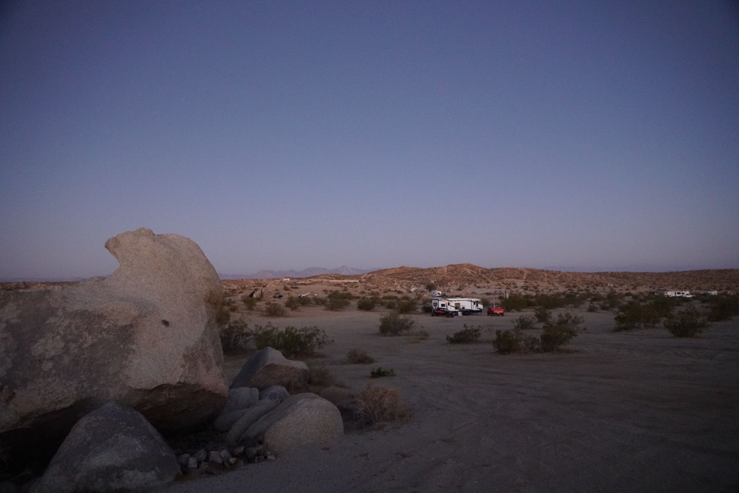 Desert landscape at dusk with large boulders and parked vehicles in the background.