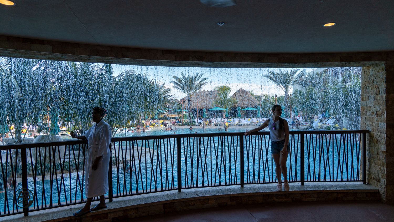 Two guests in robes stand on curved indoor balcony overlooking tropical resort pool area.