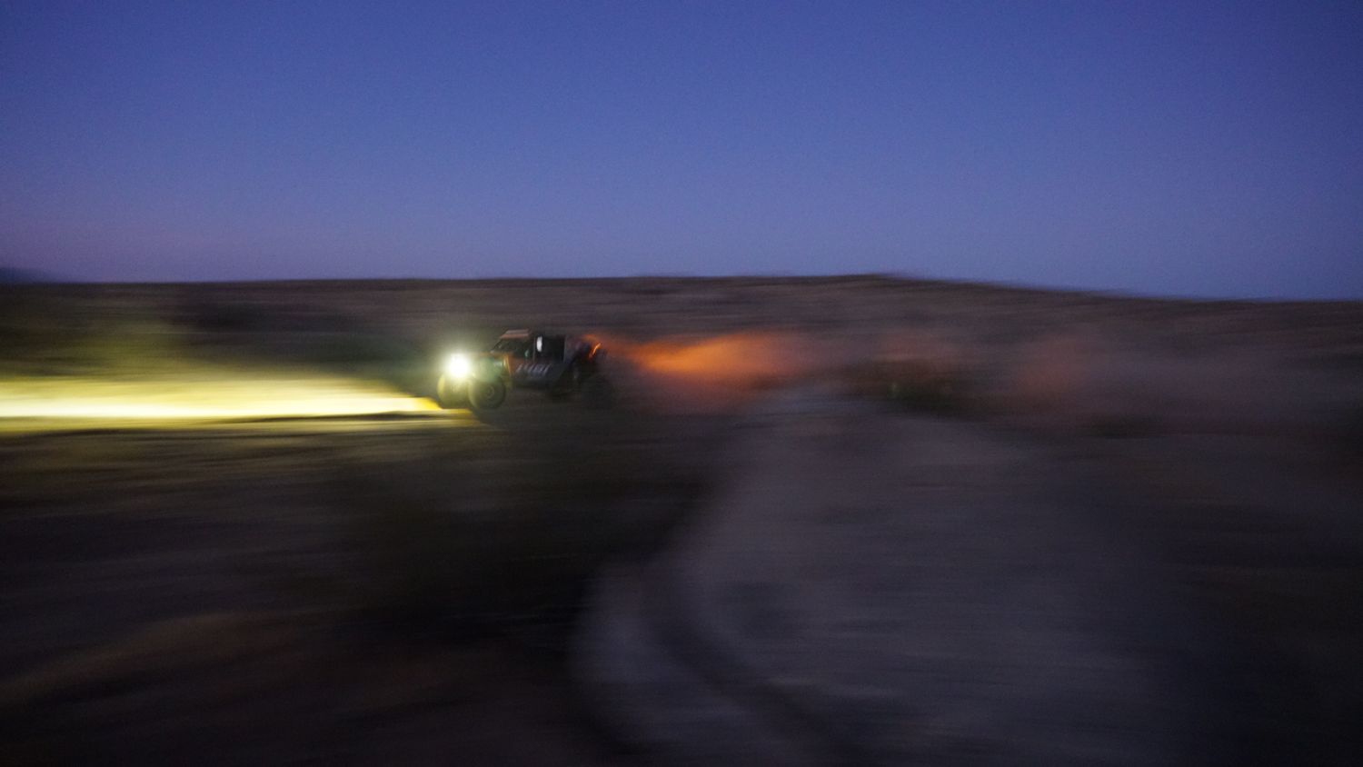 Off-road vehicle with bright headlights speeding through a dark desert landscape at dusk.