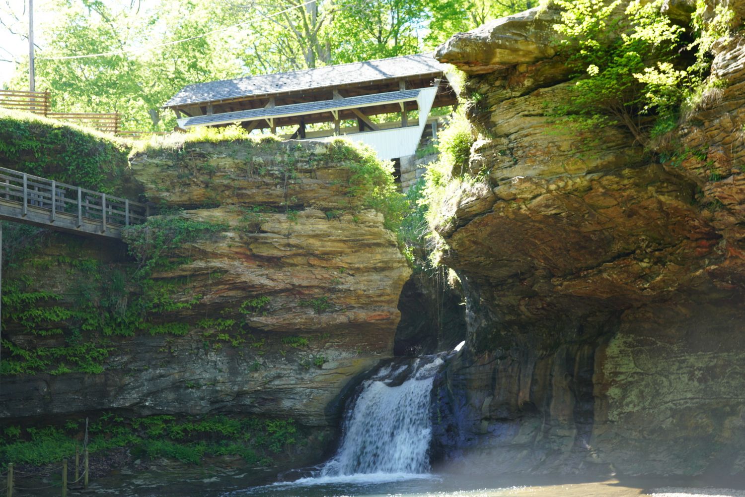 A scenic waterfall cascades down sandstone cliffs beside a covered bridge walkway surrounded by lush green trees.