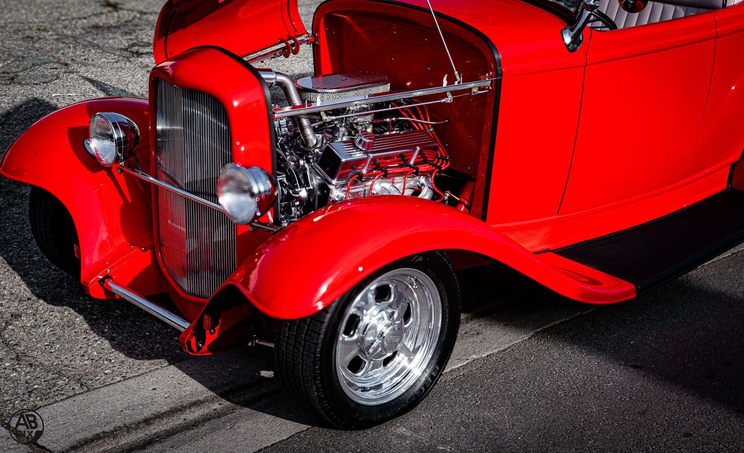 Bright red classic hot rod with exposed supercharged V8 engine and chrome details at a car show.