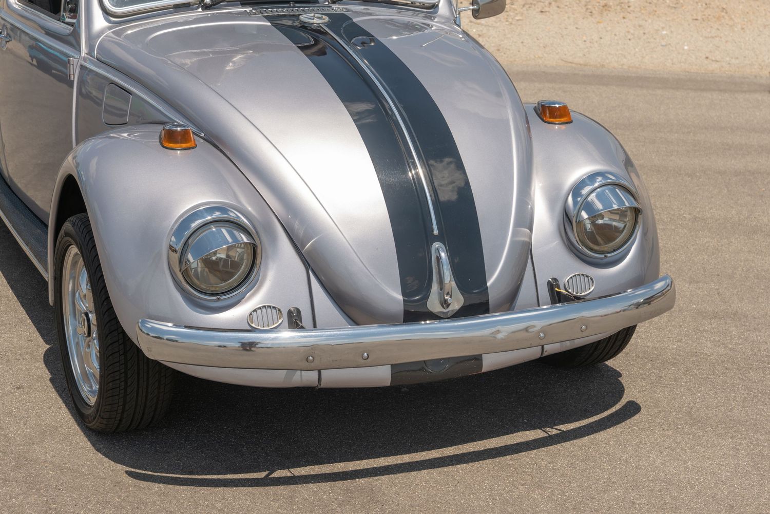 Silver vintage VW Beetle with dark racing stripes on hood, classic chrome details, parked on sunny pavement.