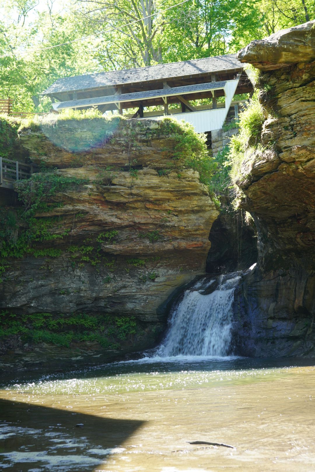 A scenic waterfall cascading over layered sandstone cliffs beneath a covered bridge surrounded by lush green trees.
