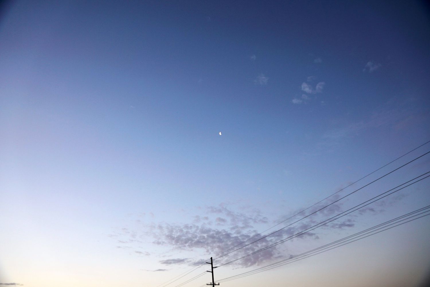 A crescent moon visible in a clear blue dusk sky above power lines and wispy clouds at twilight.