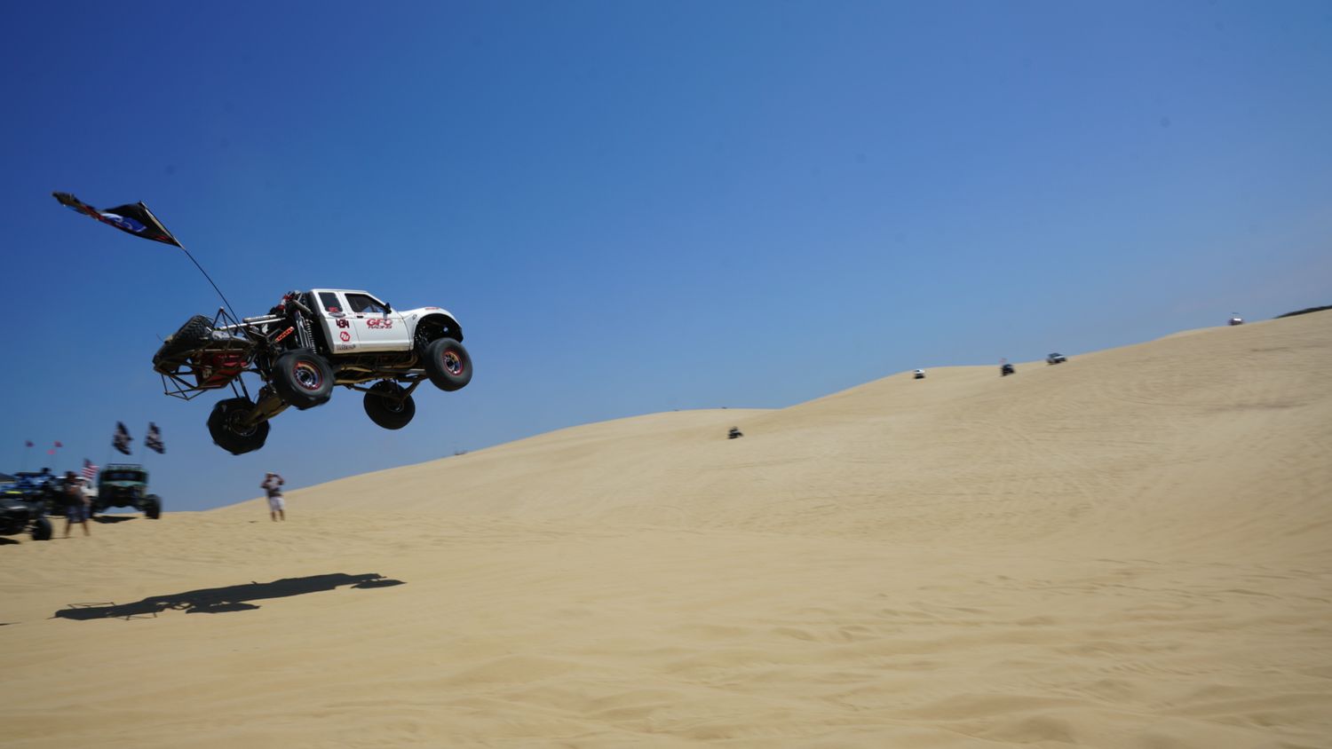 A white off-road trophy truck jumping high over sand dunes with spectators watching at a motorsport event.