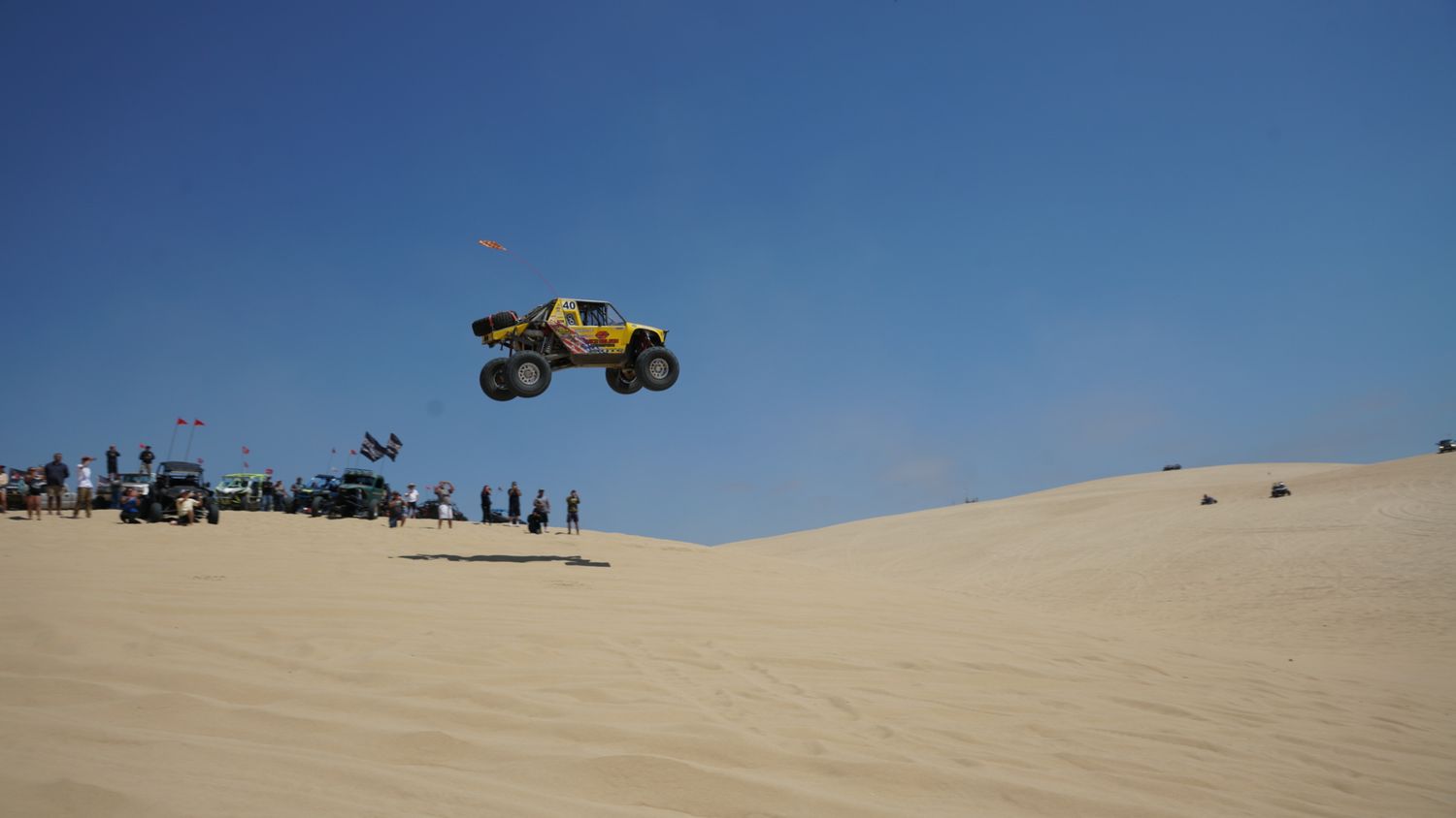 Yellow off-road buggy performing a dramatic jump over desert sand dunes with spectators watching below.