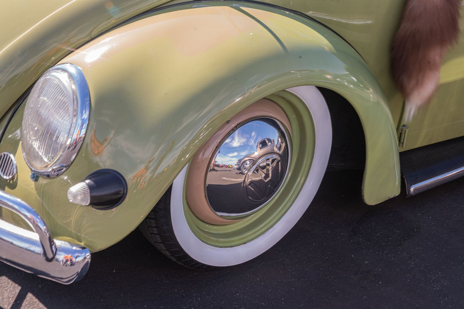 Close-up of a classic yellow VW Beetle wheel with chrome hubcap and white wall tire at a car show.