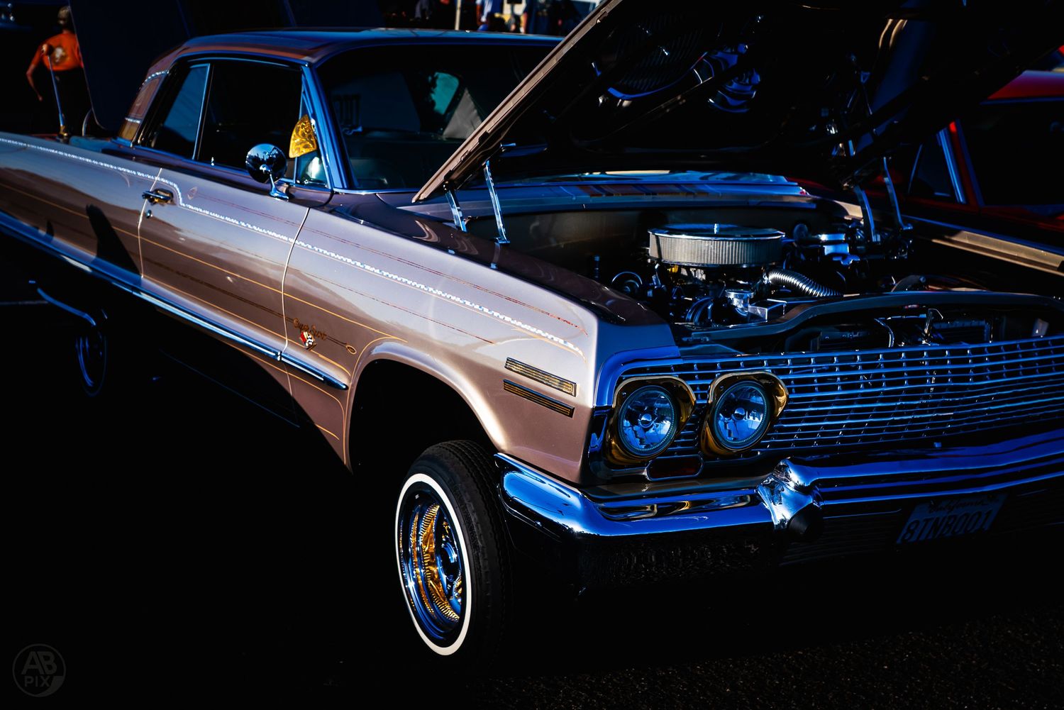 A classic lowrider Chevrolet Impala with white interior and whitewall tires illuminated at night.