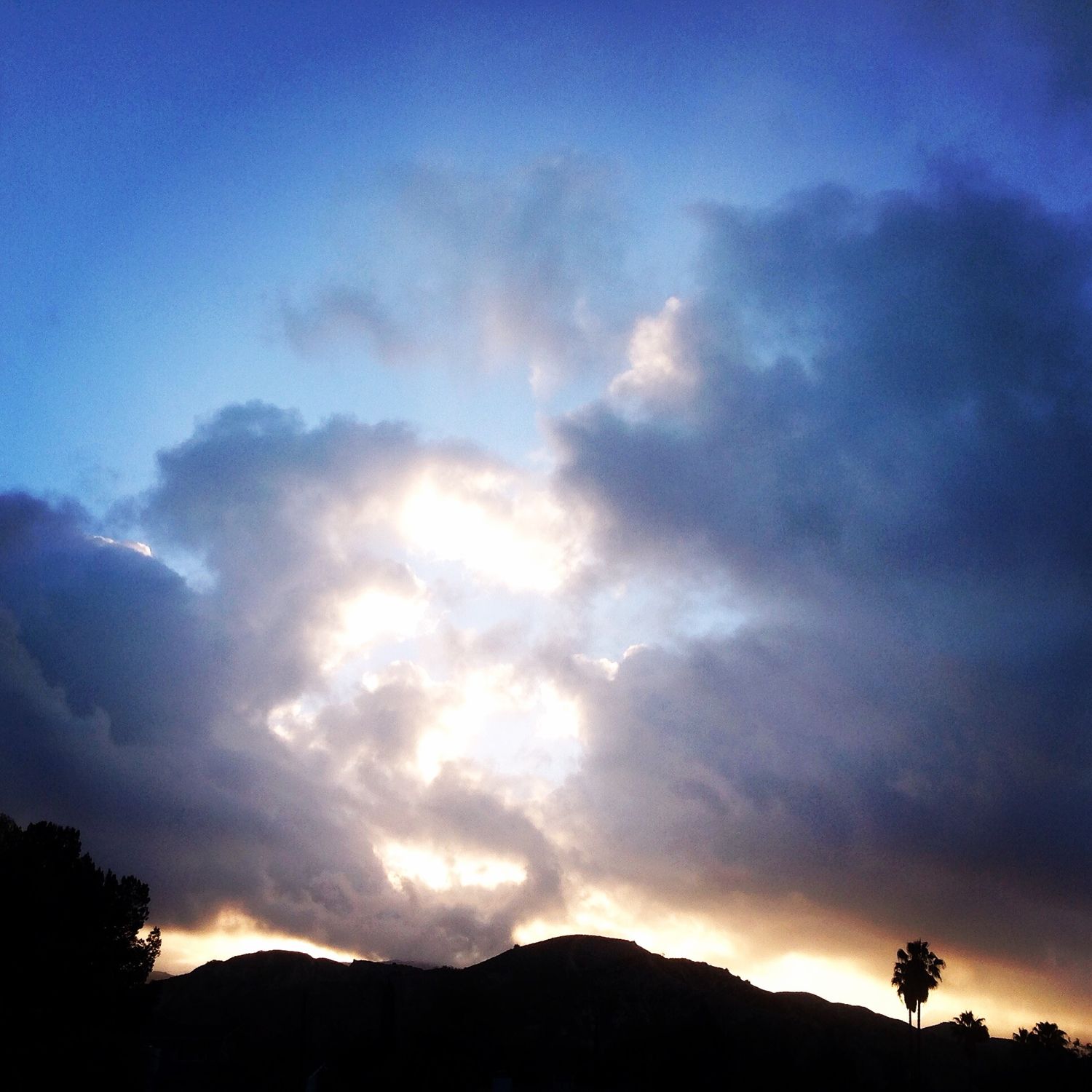 Dramatic blue sky with large glowing clouds backlit by sunset over a mountain silhouette.