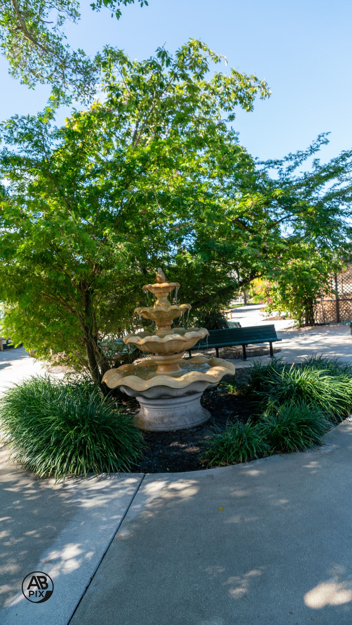 A three-tiered stone garden fountain surrounded by lush green trees and landscaped plants outdoors.