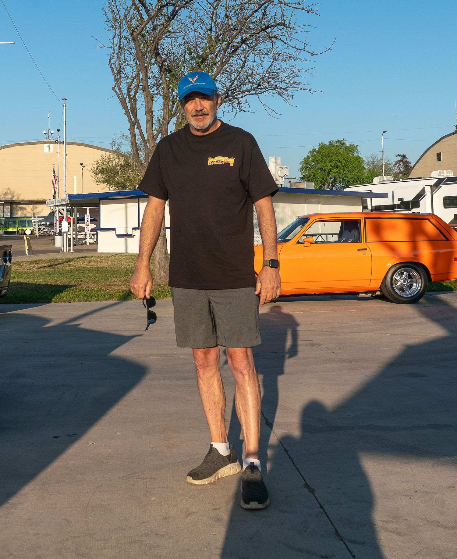Man in blue cap and black Thrasher tee walks in parking lot beside a classic orange muscle car on a sunny day.