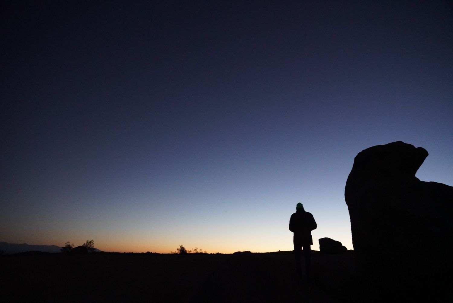 Person standing beside large rock silhouetted against vivid desert sunset sky.