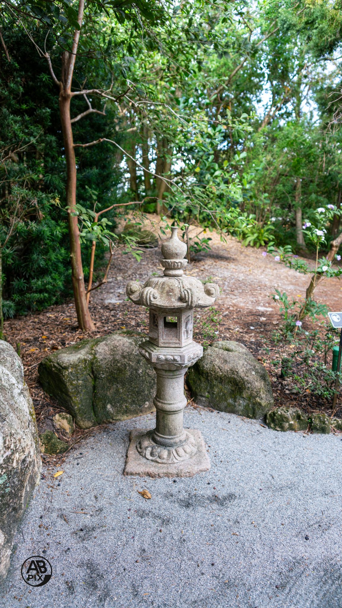 Ornate stone Japanese lantern on gravel path surrounded by rocks and lush green trees.