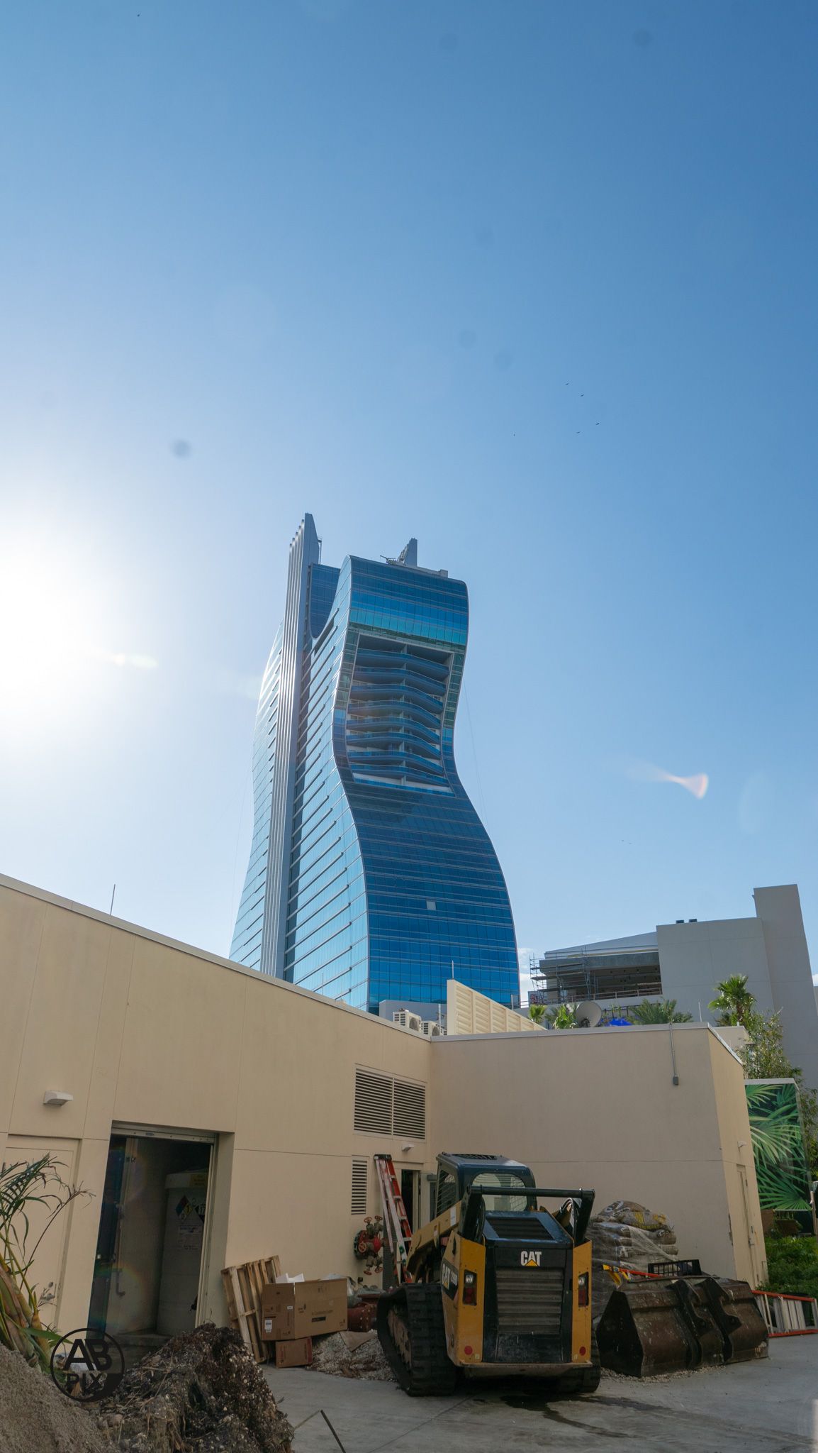 Curved blue glass skyscraper with CAT construction equipment in foreground under sunny sky.