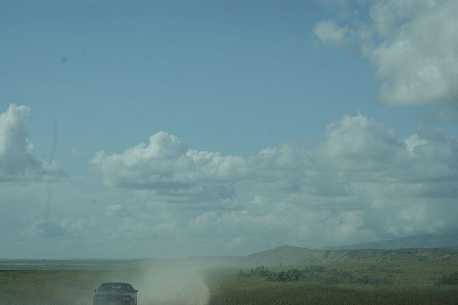 Dusty dirt road with vehicle kicking up dust under blue sky with white clouds.
