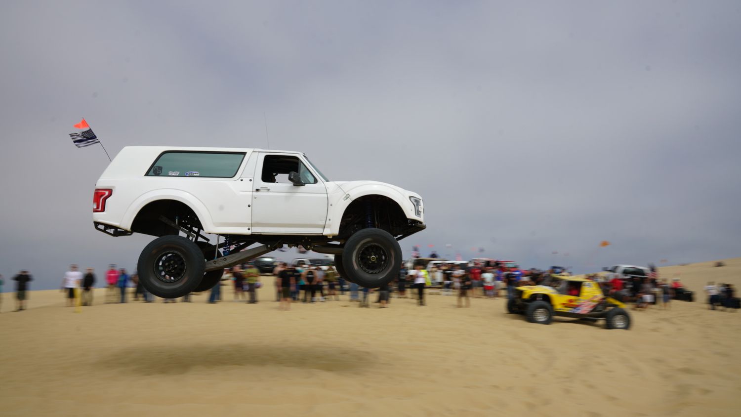 White lifted truck catches air over sand dunes at off-road desert racing event with crowd watching.