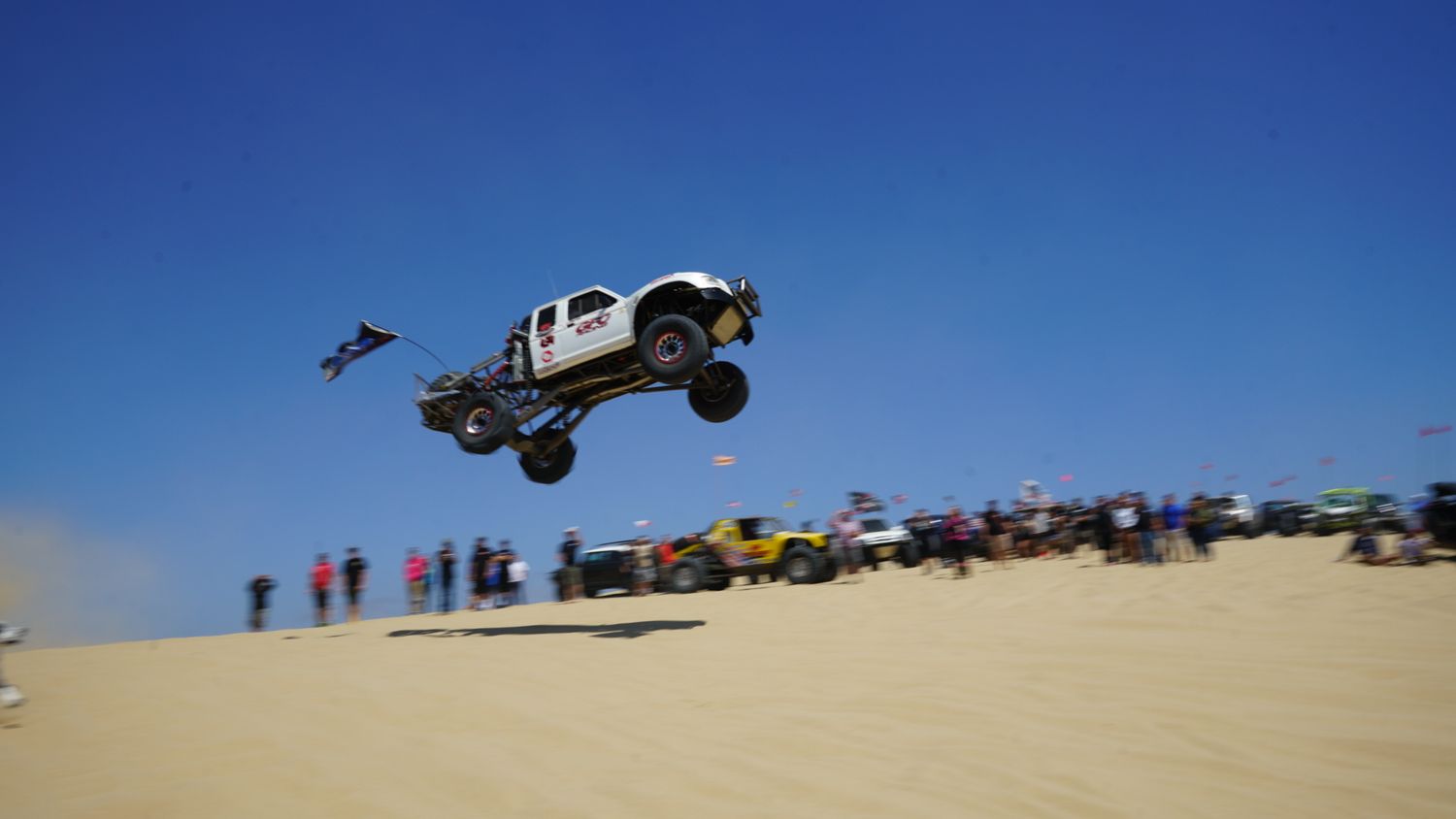 A white off-road trophy truck catching big air over sand dunes at an outdoor motorsport competition.