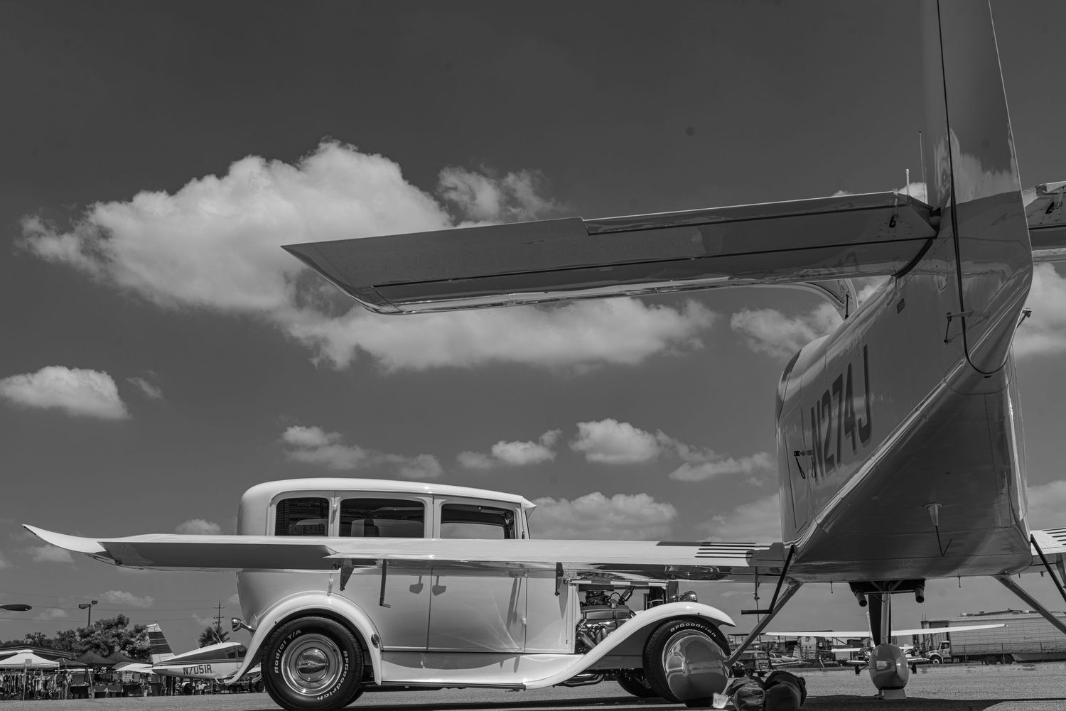 Black and white photo of a classic hot rod car parked beside a vintage airplane on a sunny airfield.