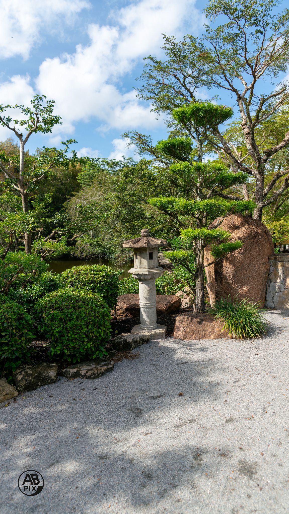 Stone lantern in a Japanese garden with manicured pine trees, lush shrubs, and gravel path under blue sky.