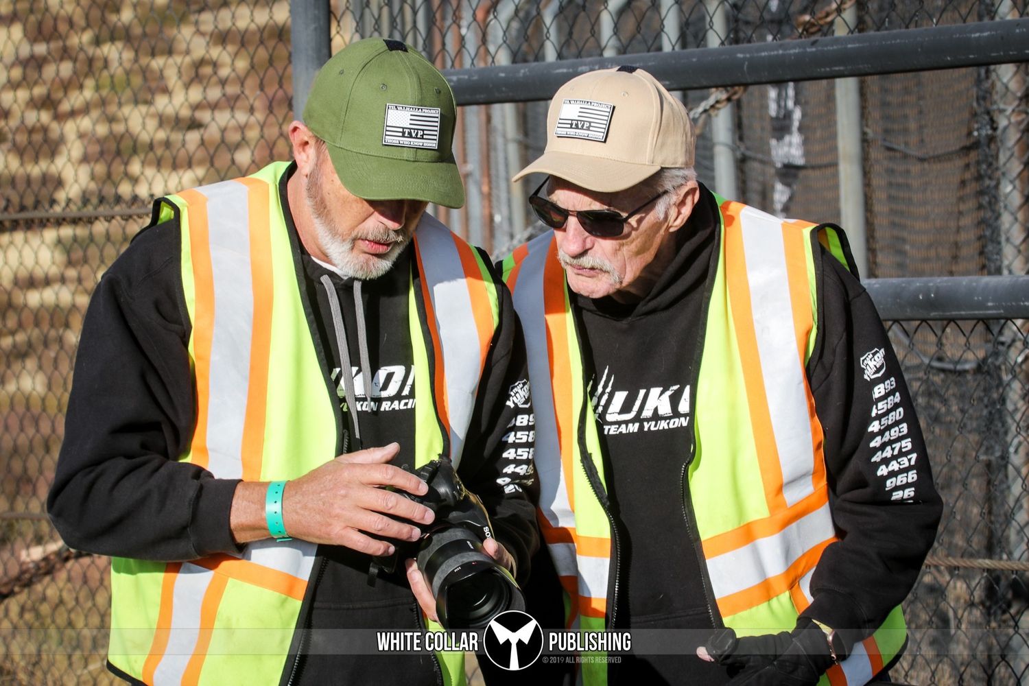 Two photographers in safety vests and caps review camera shots outdoors at an event.
