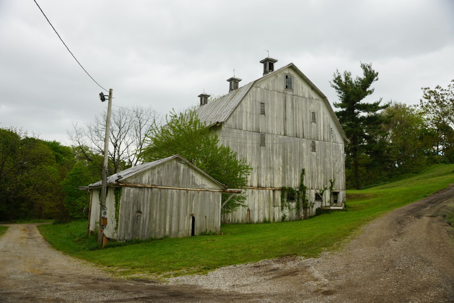 Weathered white wooden barn with gambrel roof and small outbuilding along a rural gravel driveway.
