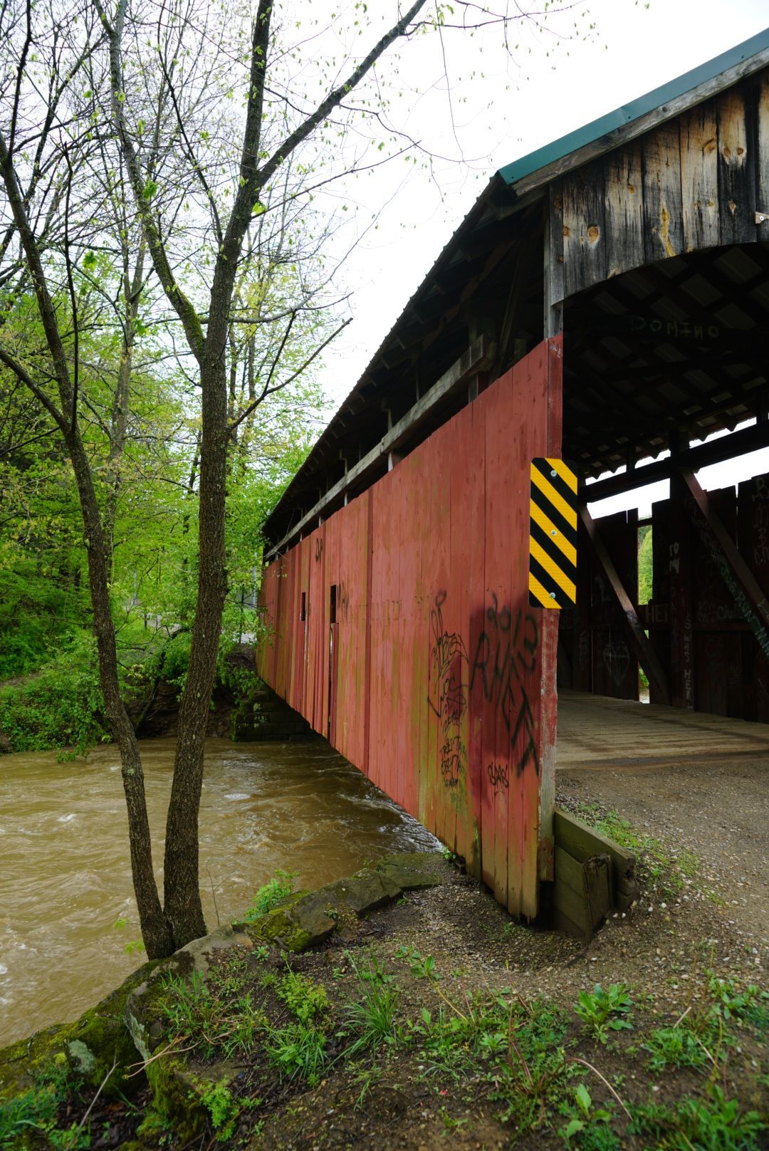 A red covered bridge with yellow caution stripes sits above a rushing muddy creek.