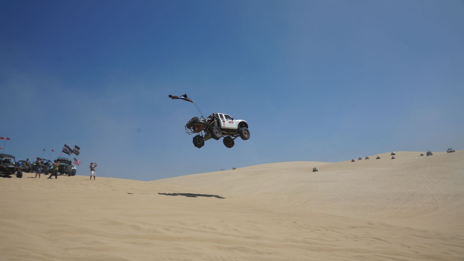 White off-road truck launches into the air over sandy dunes at a desert motorsport event.