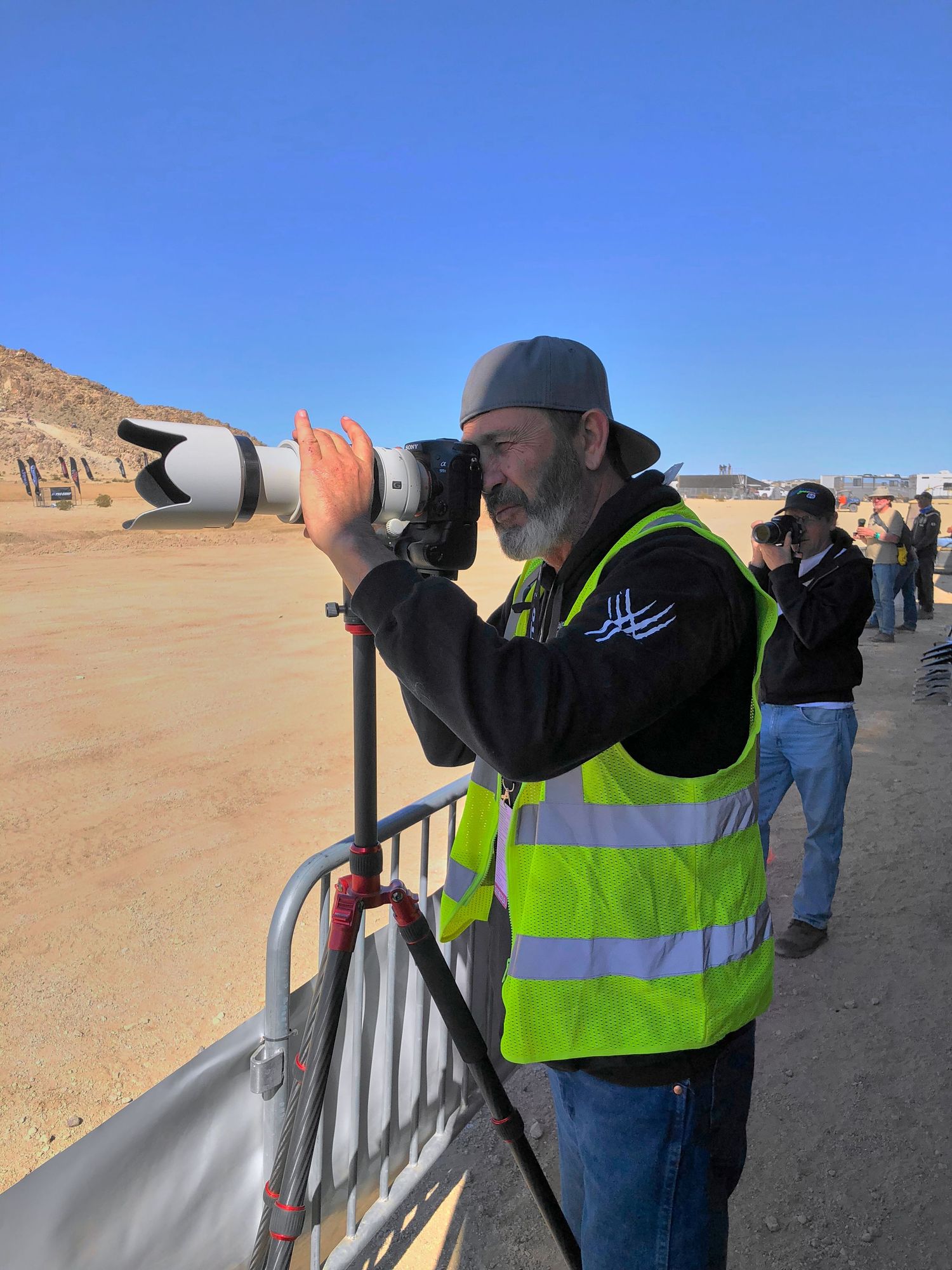 Photographer in yellow safety vest using a telephoto lens on a tripod at an outdoor event.