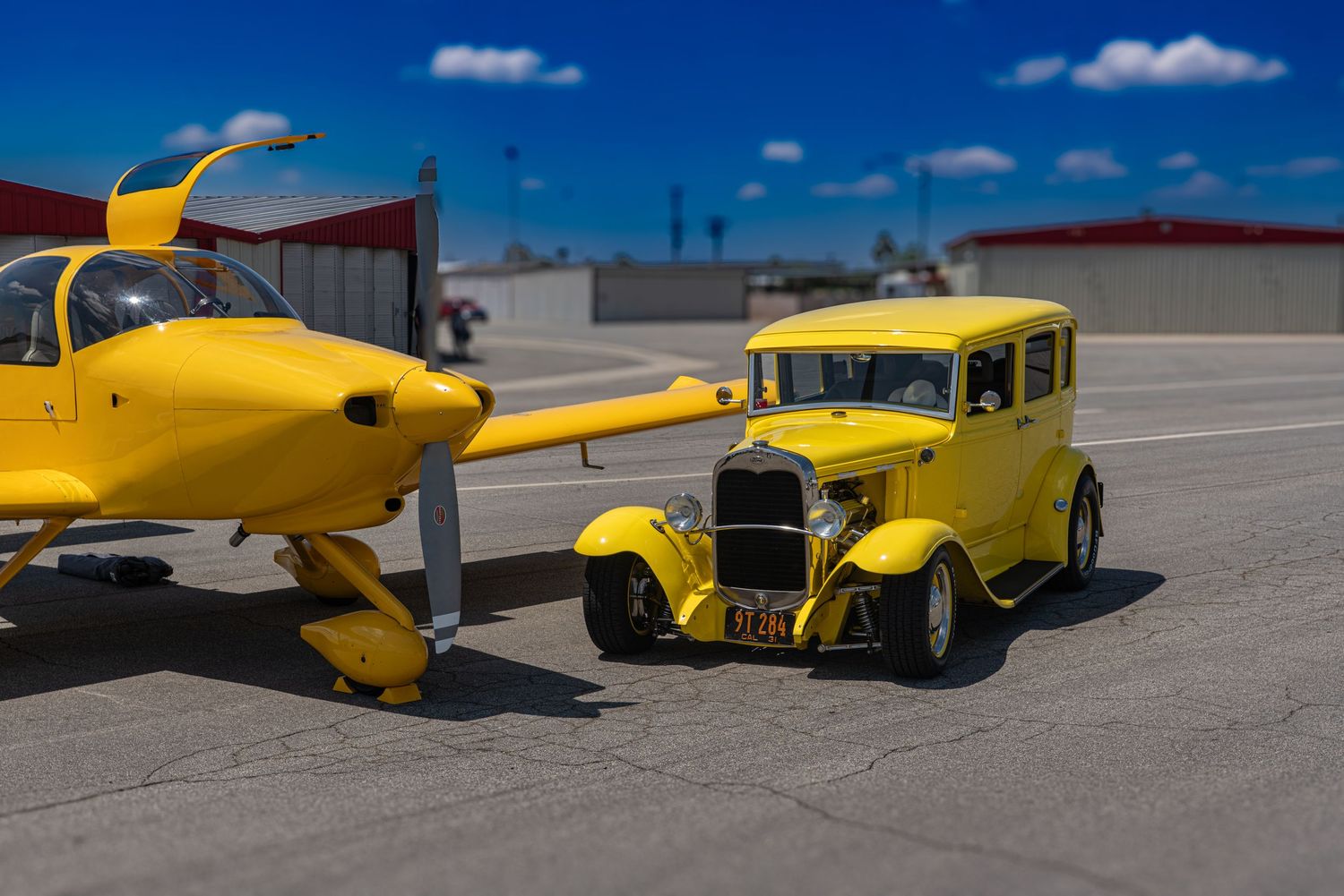 Yellow vintage hot rod car positioned next to a matching yellow small aircraft on a sunny tarmac.