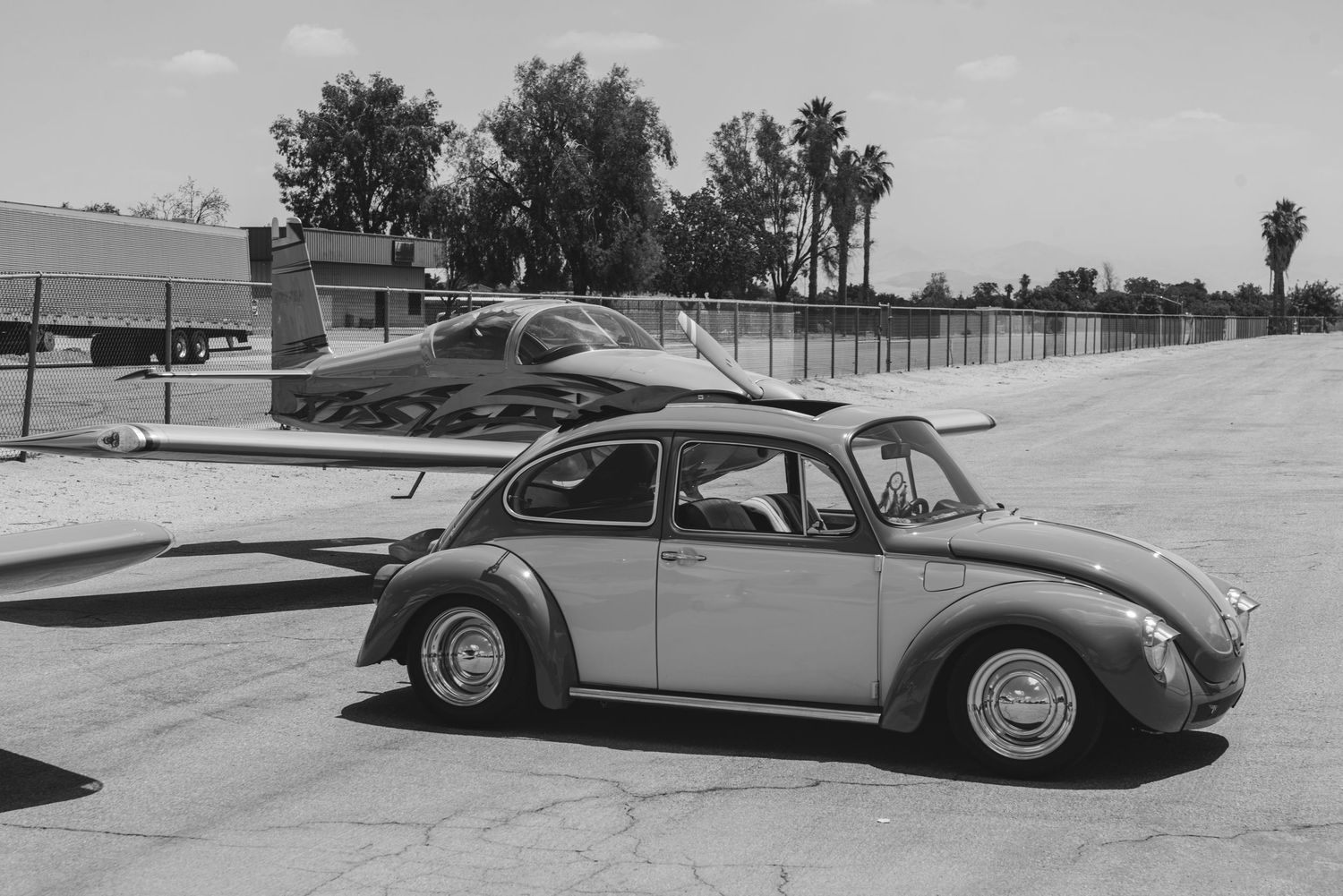 Black and white photo of a custom VW Beetle parked beside a small aircraft on an airport tarmac.