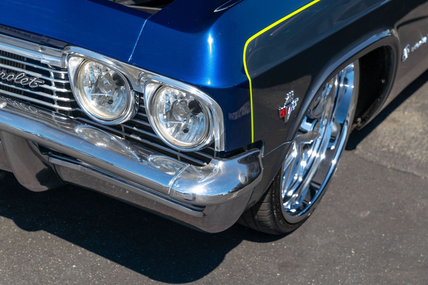 Front corner detail of a custom blue Chevrolet Impala with chrome wheels and yellow accent stripe at a car show.