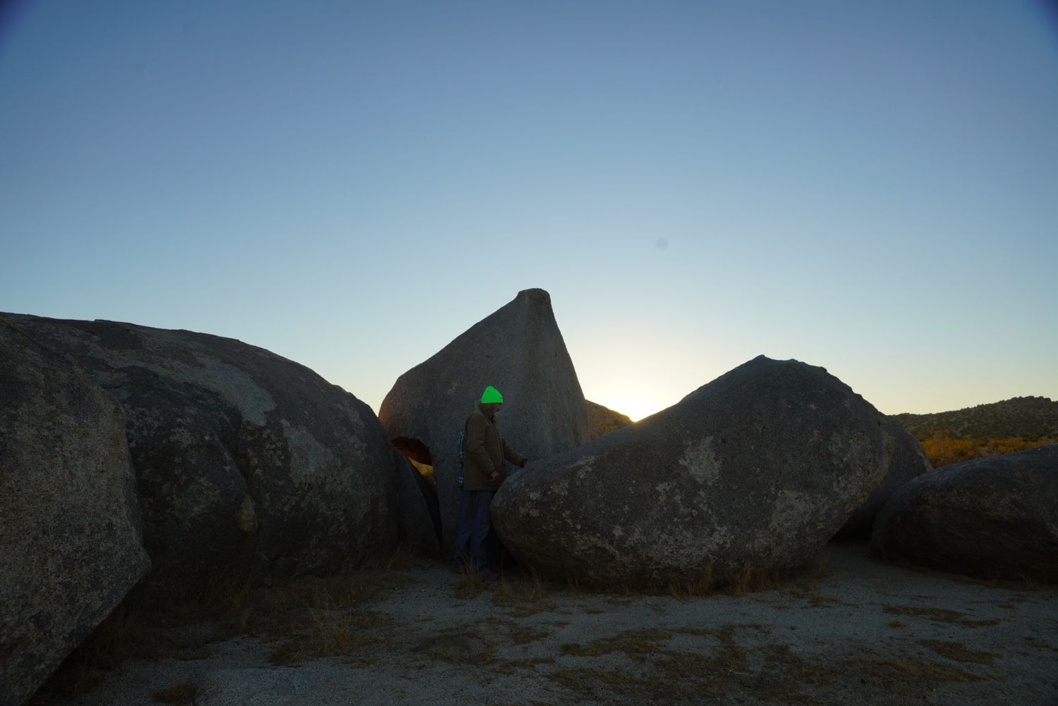 A silhouetted person stands among large granite boulders at sunset in a desert landscape.