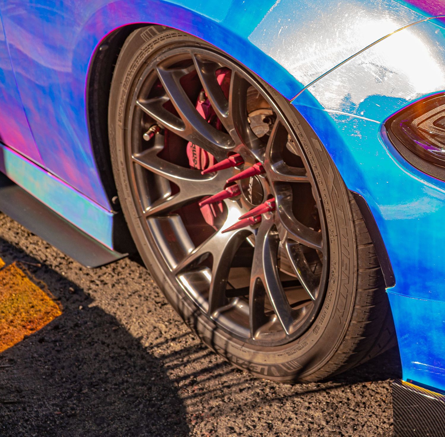 Close-up of a dark alloy wheel with red brake calipers on a holographic chrome wrapped sports car.