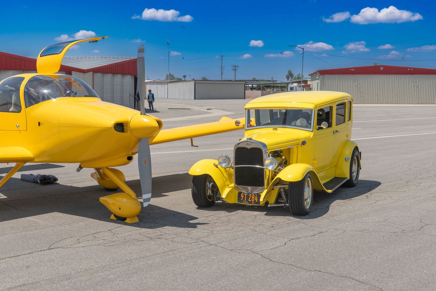 Yellow vintage hot rod car parked next to a matching yellow small aircraft on a sunny airport tarmac.