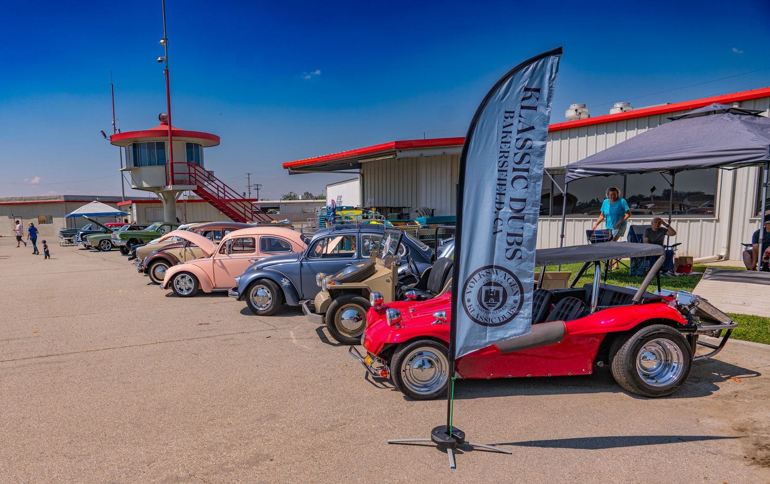 Classic VW Beetles and vintage cars on display at an outdoor car show near a red control tower on a sunny day.