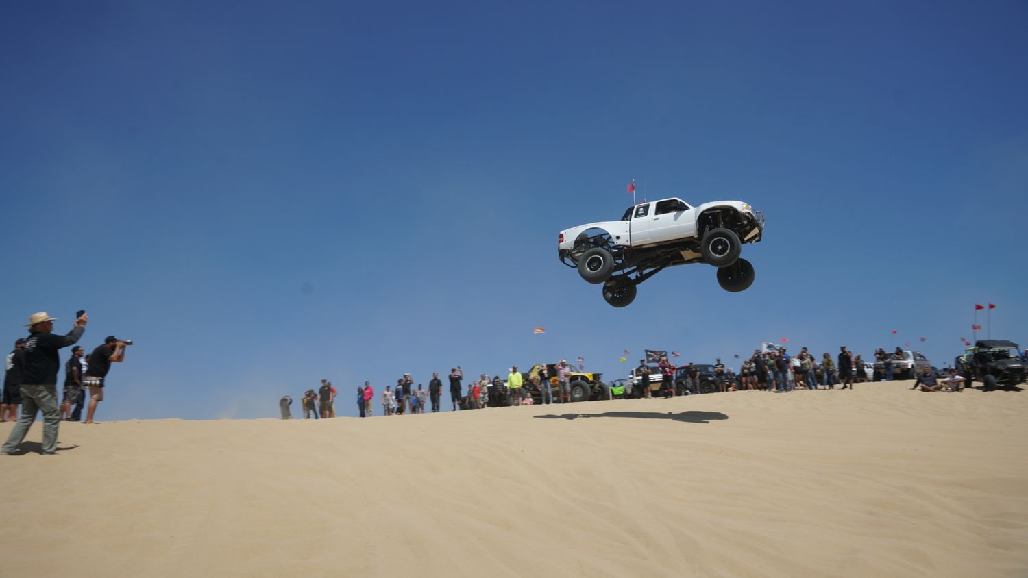 White trophy truck soars high above sand dunes while spectators watch the off-road jump competition.