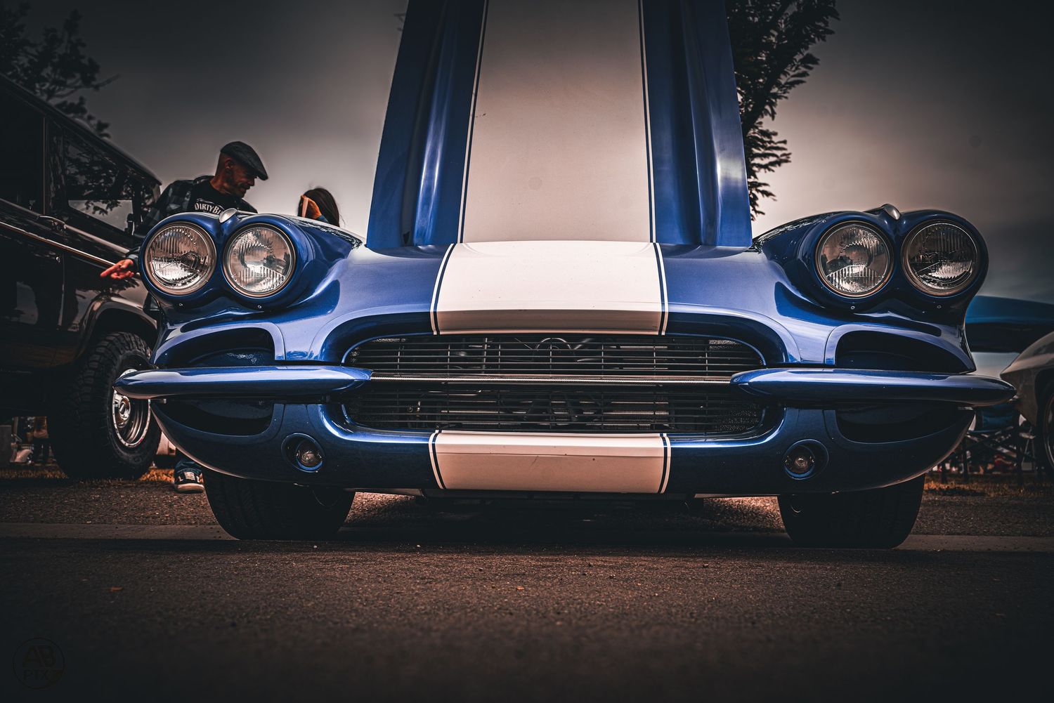 Blue classic Corvette with white racing stripes at a car show, low angle front view.