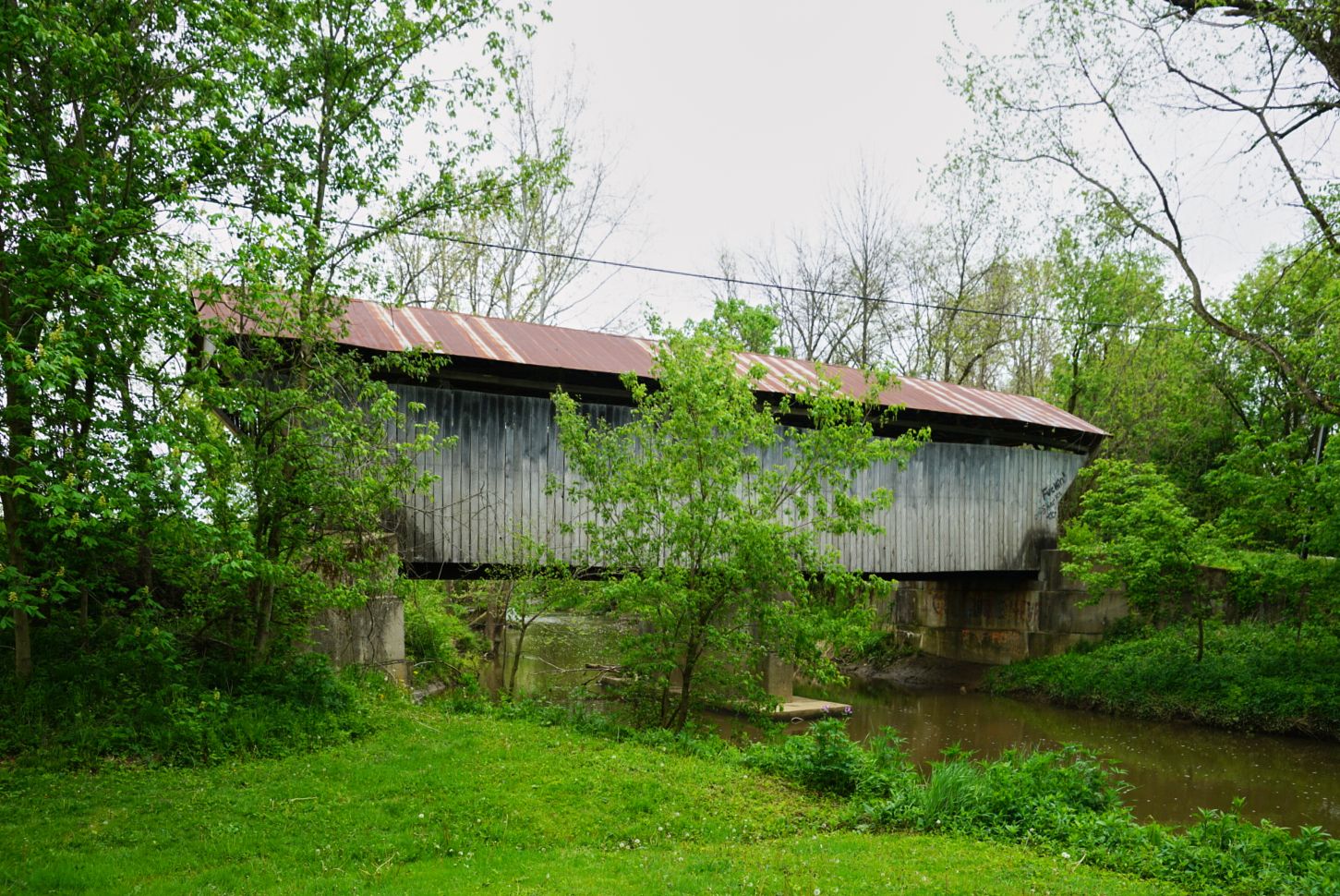 Rustic historic covered bridge with corrugated metal roof spanning a calm creek surrounded by lush spring green trees.