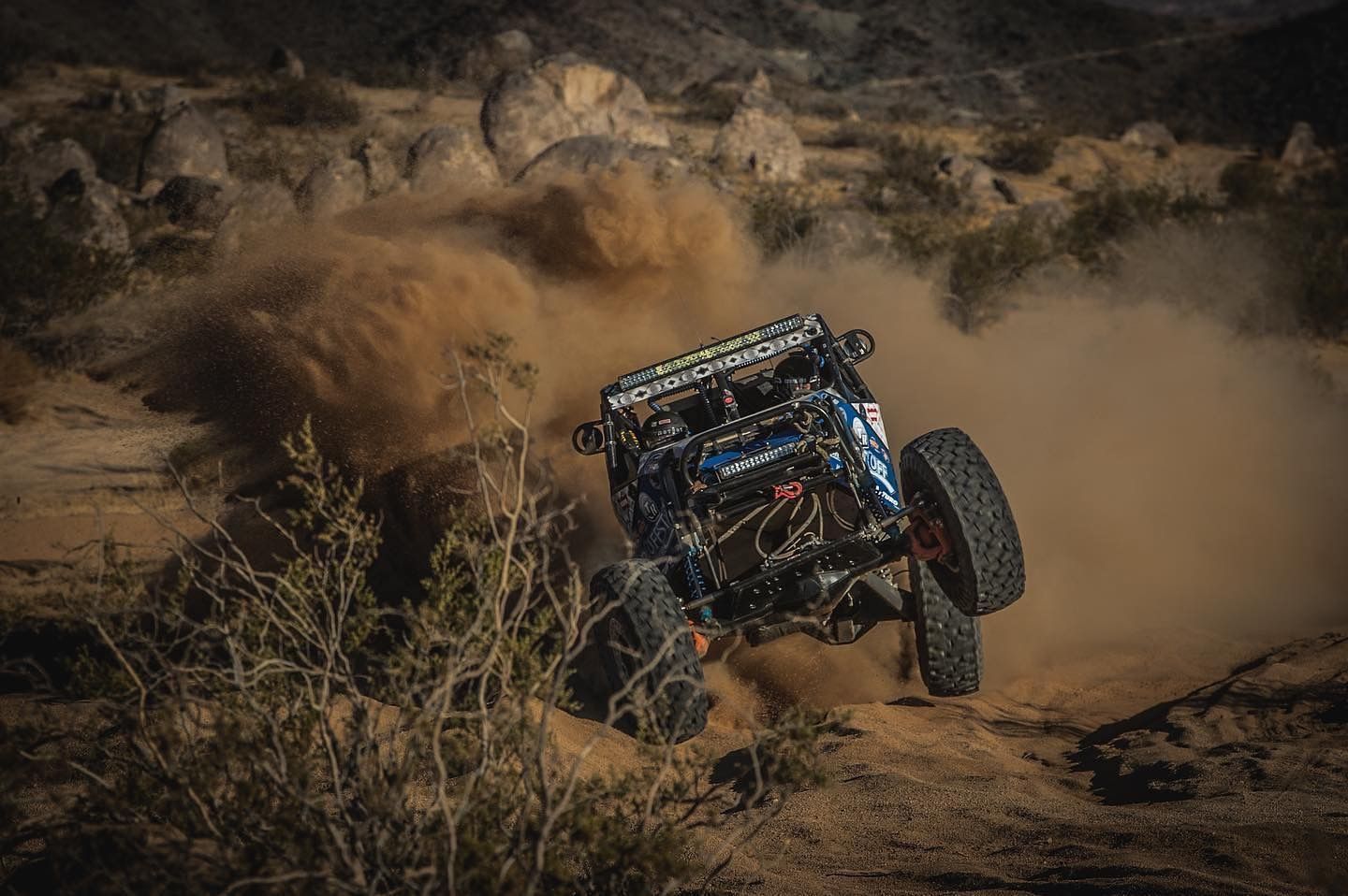 Blue off-road buggy climbing rocky desert terrain with dramatic dust cloud.