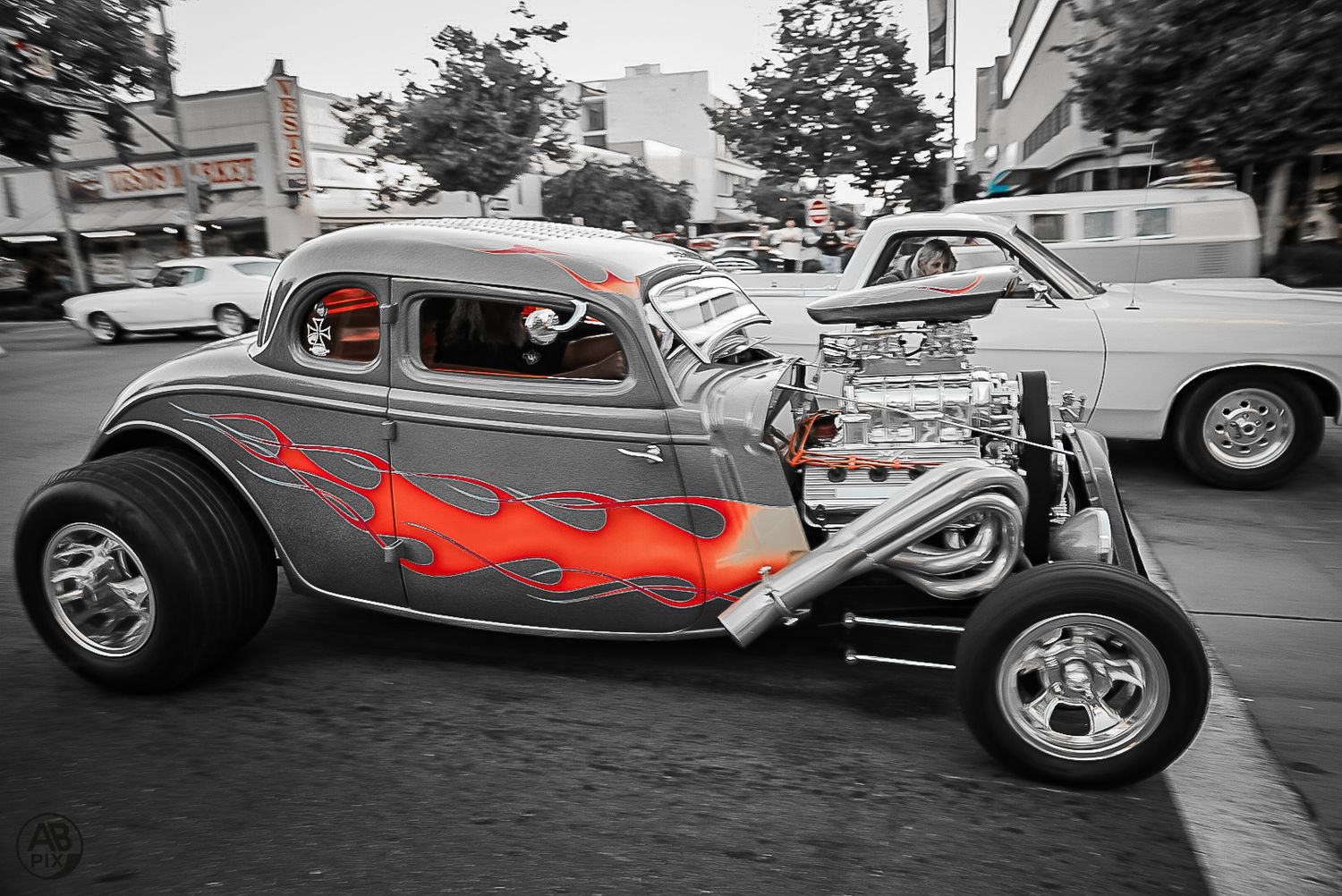 Vintage hot rod with red flames and exposed chrome engine driving on a city street.